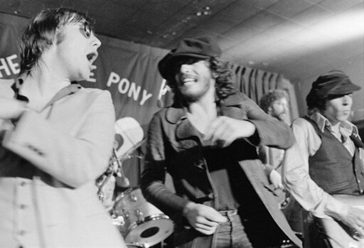 Four men energetically perform on stage, dressed in 1970s fashion. One sings passionately, while others smile under dim club lighting. Energetic atmosphere.