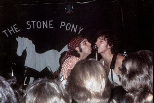 Musicians passionately sing into a shared microphone on stage at The Stone Pony. The crowd is engaged, and a backdrop features a white horse silhouette.