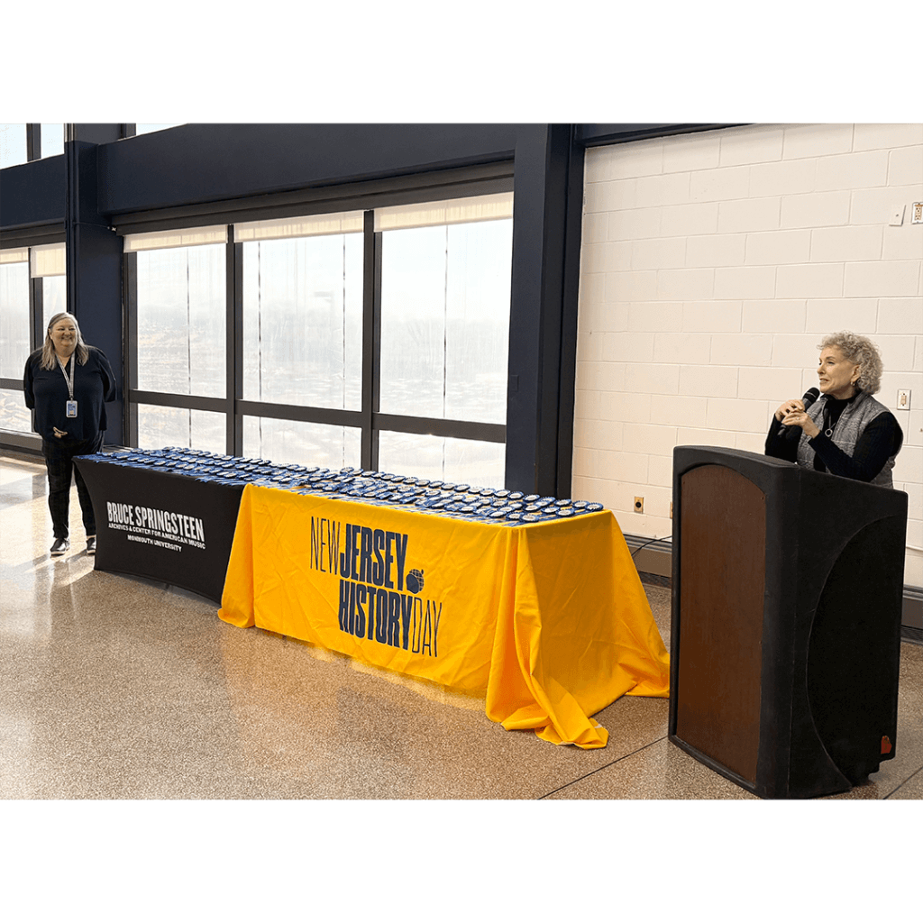 A speaker at a podium addresses an audience in a bright room. Two tables are covered with medals, draped in "BRUCE SPRINGSTEEN" and "NEW JERSEY HISTORY DAY" cloths.
