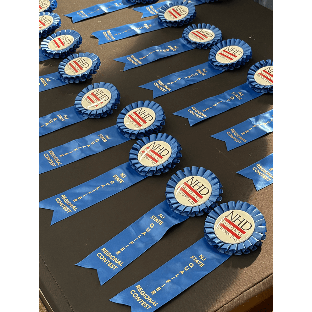 Blue ribbon awards are neatly arranged in rows on a table. They feature rosettes labeled "NHD National History Day NJ State Regional Contest," conveying achievement.