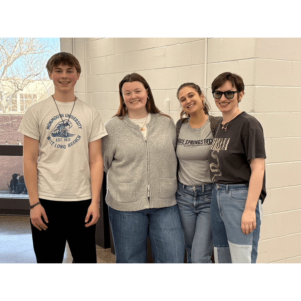 Four people stand close together smiling in a casual setting with bright light. They wear jeans and printed shirts, conveying a cheerful, relaxed vibe.