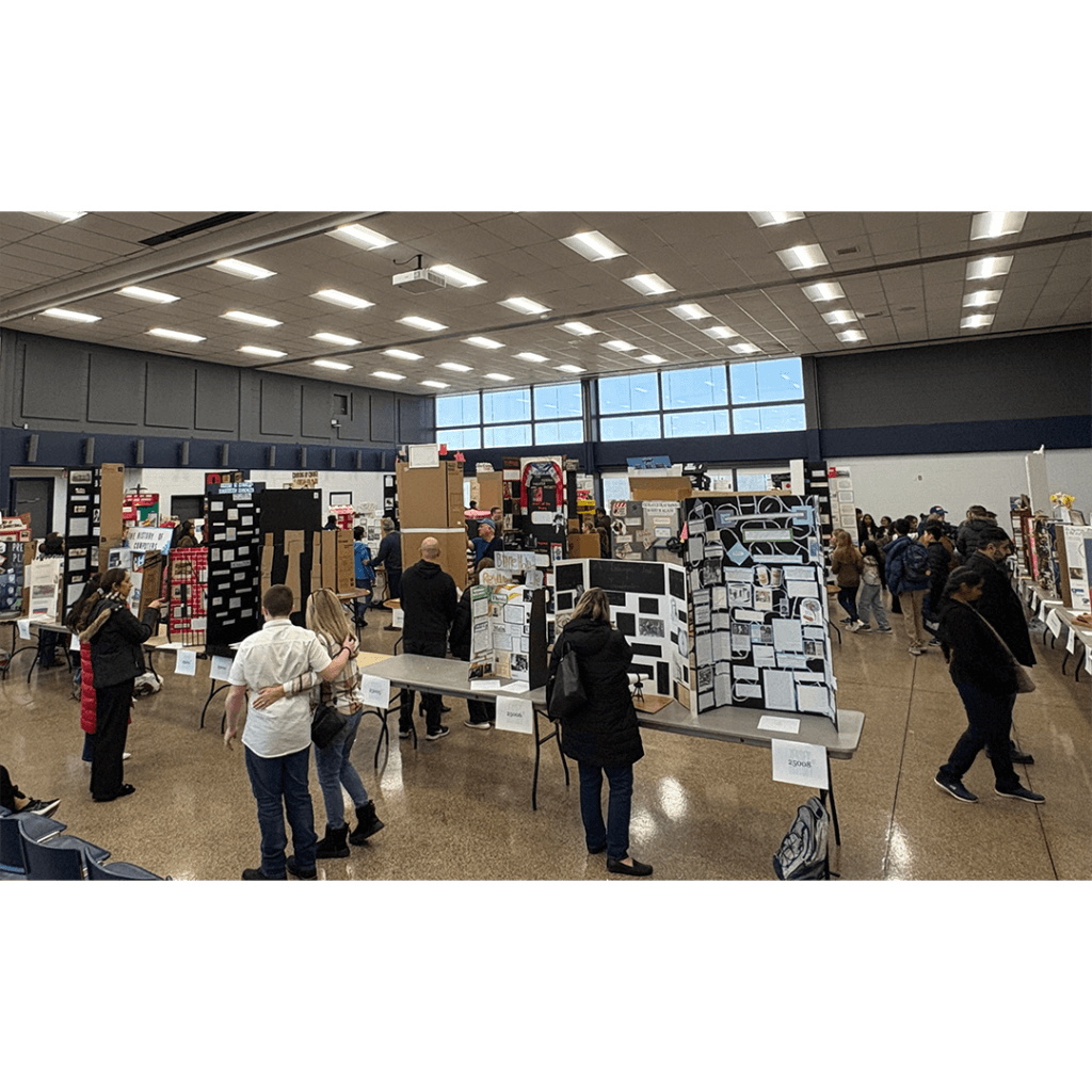 A spacious room with high ceilings hosts a science fair. Numerous display boards, arranged on tables, showcase projects. People of all ages visit exhibits, creating a lively, educational atmosphere.