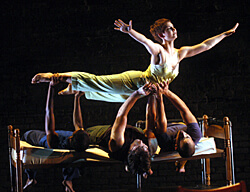A graceful dancer in a flowing dress is elegantly held aloft by three performers lying on a bed, set against a dark stage, exuding strength and harmony.