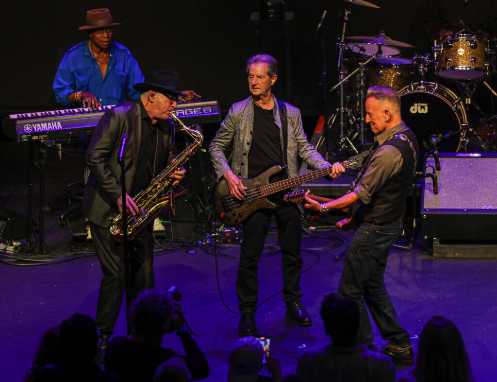 Musicians energetically perform on stage; a saxophonist in a hat, a bassist in gray jacket, and a guitarist interact, with a keyboardist in blue behind them.