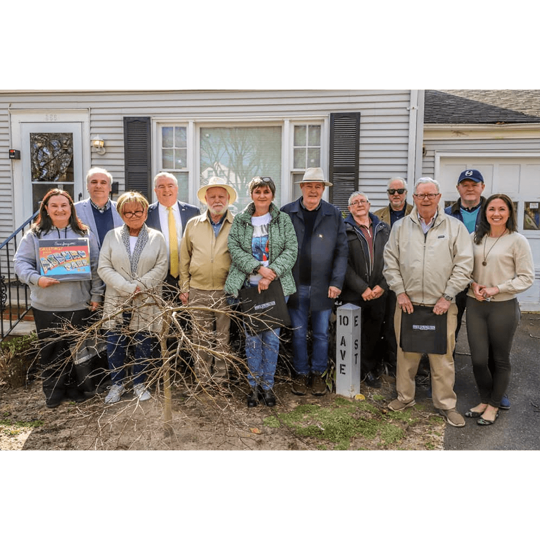 A group of twelve adults stand smiling in front of a house. They are casually dressed, showing a sense of camaraderie. A small bare tree is in the foreground.
