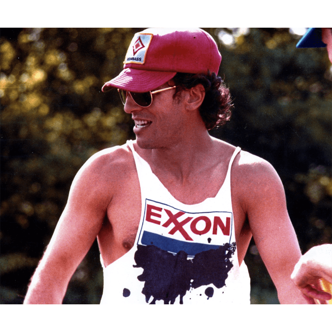 Man wearing a red cap and sunglasses, smiling outdoors. He has on a white tank top with "Exxon" in bold letters, set against a blurred natural background.