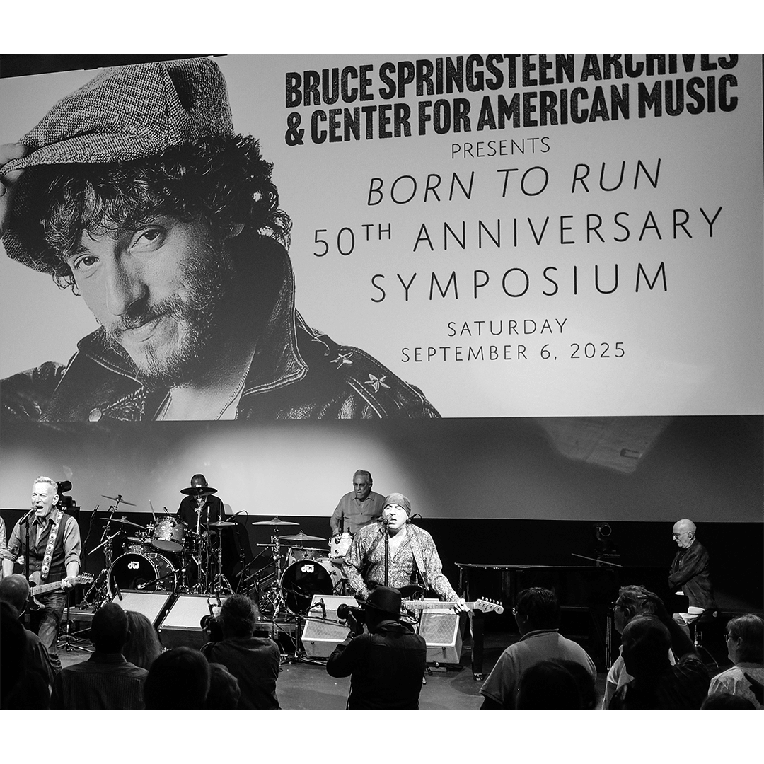 Black-and-white photo of a live band performing beneath a large screen showing Bruce Springsteen's image and "Born to Run 50th Anniversary Symposium."