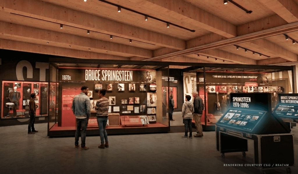 Exhibition space with Bruce Springsteen displays, featuring album covers and memorabilia from the 1970s to 1990s. Visitors observe, evoking nostalgia.