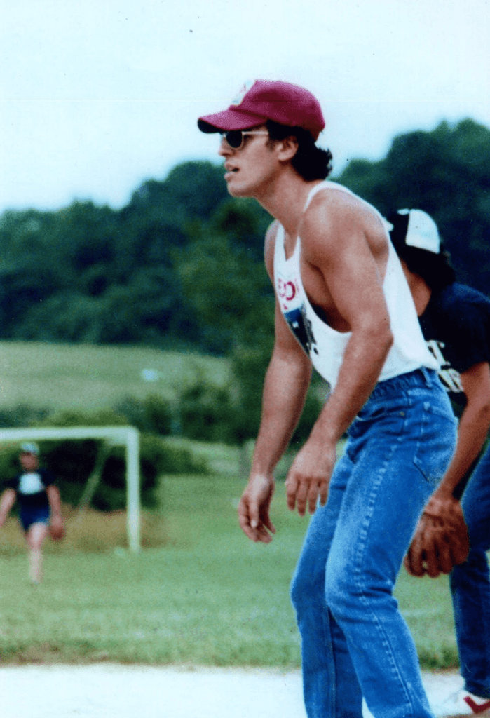 A man in a red cap and sunglasses stands in a field, wearing a white tank top and jeans. He appears focused and is leaning forward slightly.