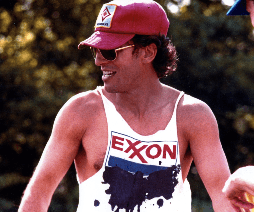 Man wearing a red cap and sunglasses, smiling outdoors. He has a white tank top with a stylized "Exxon" logo, featuring a black splash design.