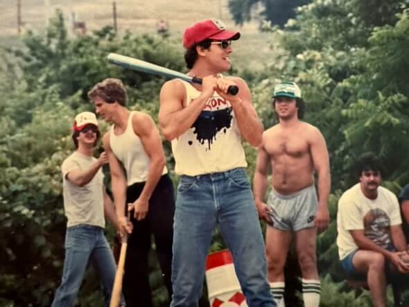 A group of young men in casual summer attire enjoying a game of baseball. The central figure holds a bat, exuding energy and confidence.
