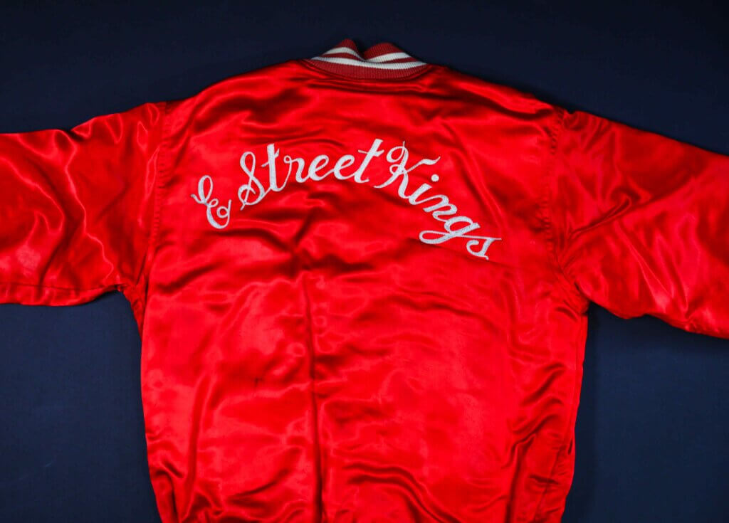 Red satin jacket with "Street Kings" embroidered in white cursive on the back. The jacket has a glossy texture, exuding a vintage, bold look.
