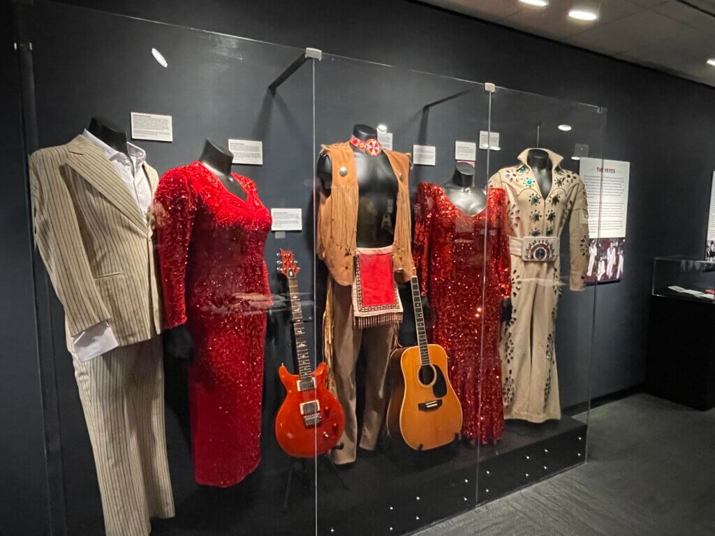 Display case with colorful vintage costumes and guitars, including two red sequined dresses, a beige fringed vest outfit, and an embroidered suit.