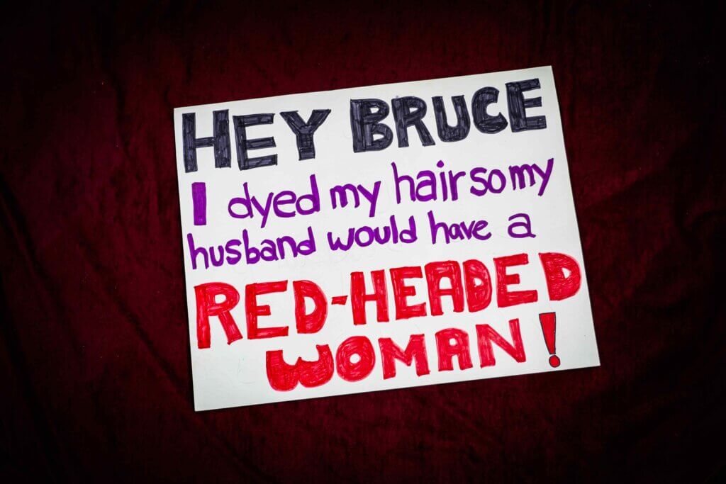 A handwritten sign on a dark red background reads, "Hey Bruce, I dyed my hair so my husband would have a red-headed woman!" in black, purple, and red letters.