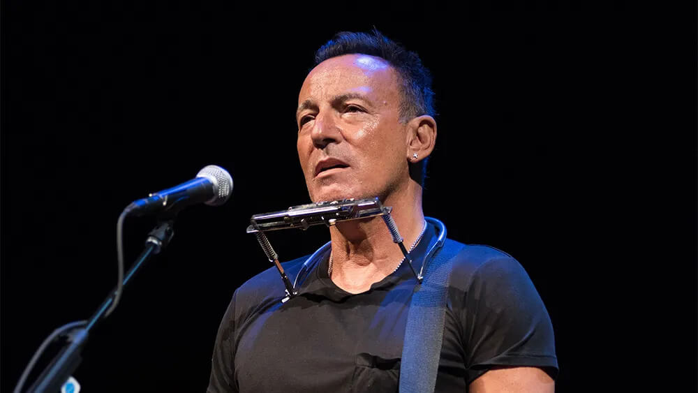 A musician stands on stage with a harmonica holder around his neck, wearing a black T-shirt. He has a focused expression, and a microphone is nearby.