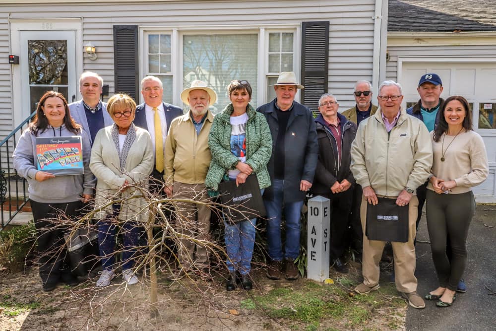 A group of twelve adults stand smiling in front of a house. They are casually dressed, showing a sense of camaraderie. A small bare tree is in the foreground.