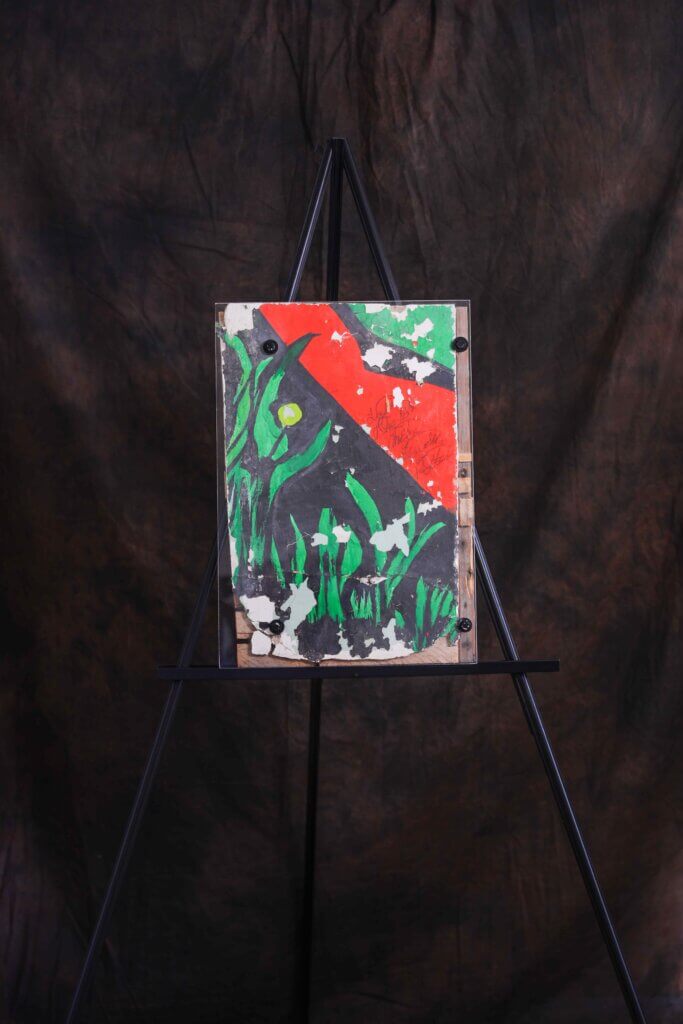 Painting on an easel with abstract strokes of green, red, and black resembling leaves, set against a dark, textured background.