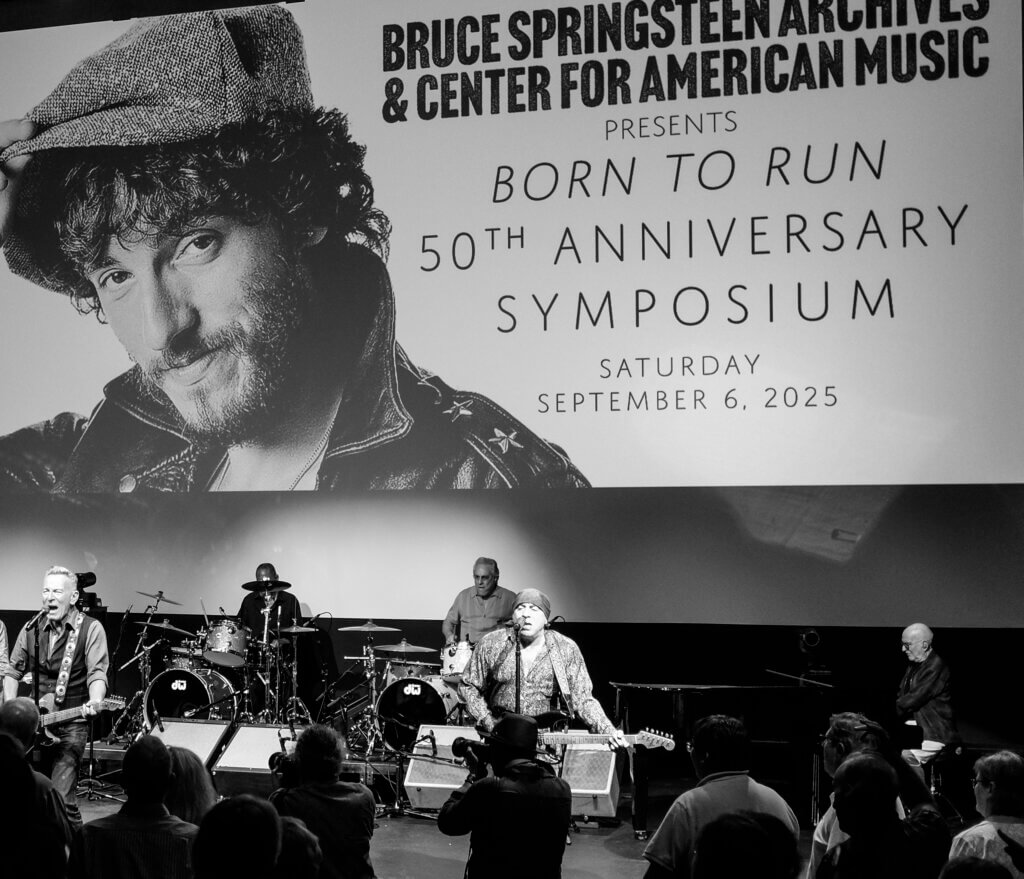 Black-and-white photo of a live band performing beneath a large screen showing Bruce Springsteen's image and "Born to Run 50th Anniversary Symposium."