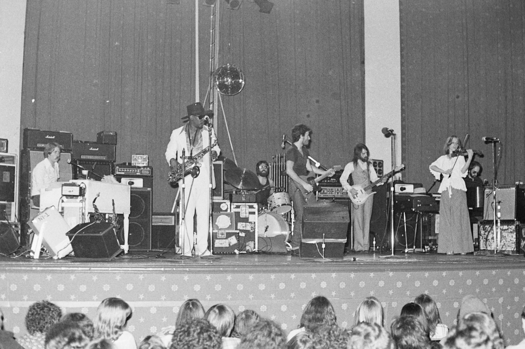 Black and white photo of a band performing on stage. Musicians play instruments including saxophone, guitar, and violin. An engaged audience watches.