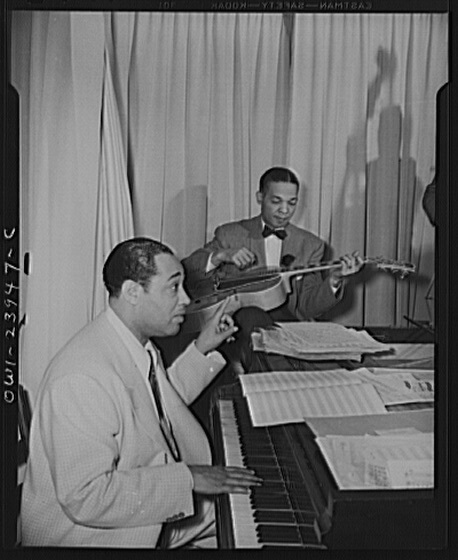 Two musicians in suits perform; one plays piano, gesturing expressively, while the other strums a guitar. Sheet music is visible, suggesting a rehearsal setting.