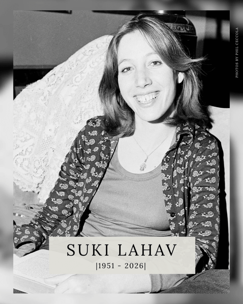 Black and white photo of a smiling woman with shoulder-length hair, wearing a patterned shirt. She is sitting indoors, and text below says "Suki Lahav, 1951-2026."