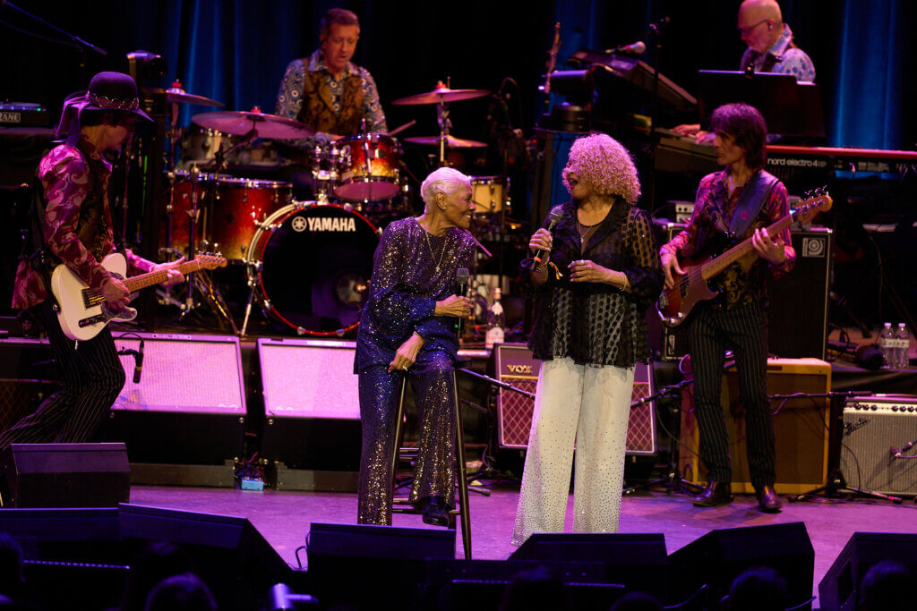 Two women sing together on stage - Darlene Love and Dionne Warwick. One seated and glittering in a sparkly suit, the other standing in a patterned blouse. Musicians play behind them, creating a lively, joyful atmosphere.