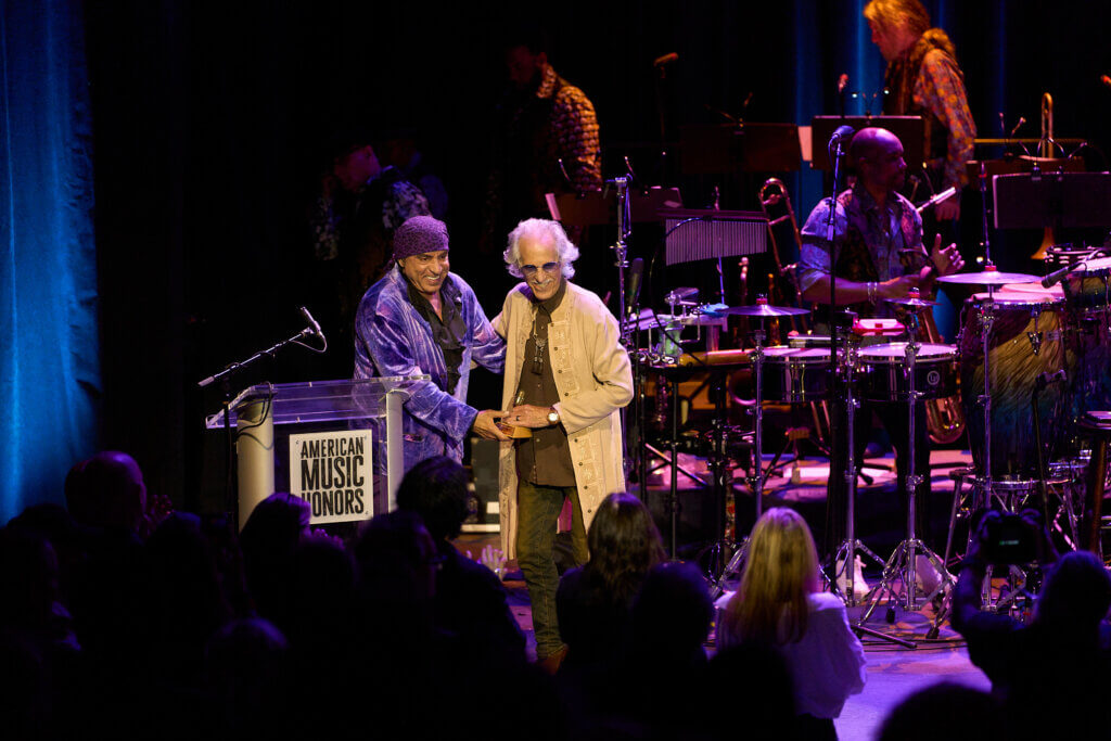 Two individuals - Stevie Van Zandt and John Densmore - stand on stage under soft lighting at the "American Music Honors" event. Musicians are visible in the background, creating a celebratory atmosphere.