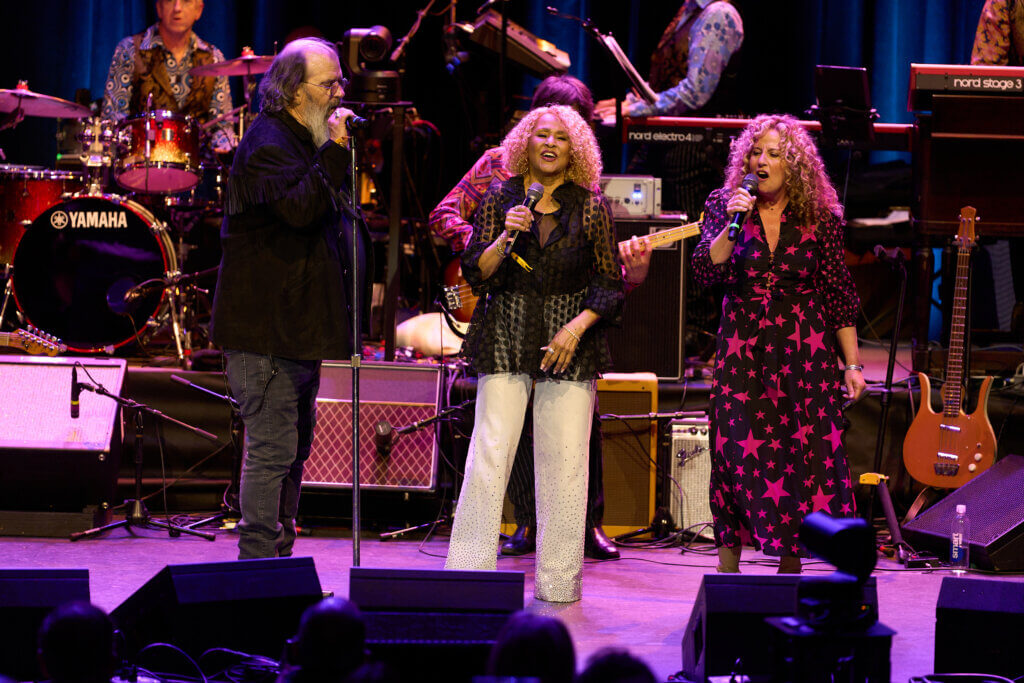 Three musicians - Steve Earle, Darlene Love and Amy Helm - energetically perform on stage, singing with microphones. A drummer and keyboardists play in the background, creating a lively atmosphere.