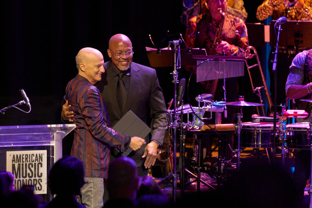 Two people - Jimmy Iovine and Dr. John - and embrace on stage at the American Music Honors. They smile warmly, surrounded by musical instruments and a podium, creating a celebratory atmosphere.