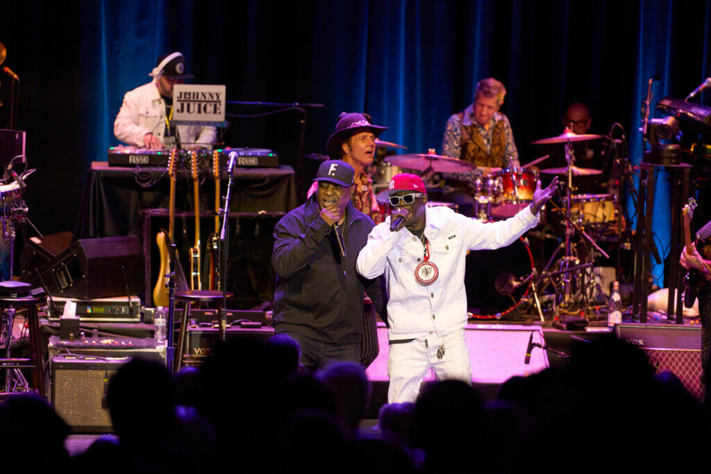 Musicians (Public Enemy) perform energetically on stage with vibrant lighting. A lively atmosphere is created by two singers in the foreground, a drummer, and a DJ in the background.