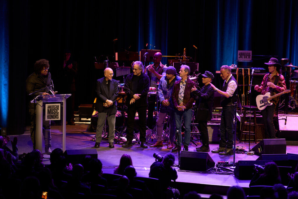 A group of musicians (The E Street Band) receives an award onstage at the American Music Honors. They stand under blue lighting, smiling and clapping, with instruments in the background.