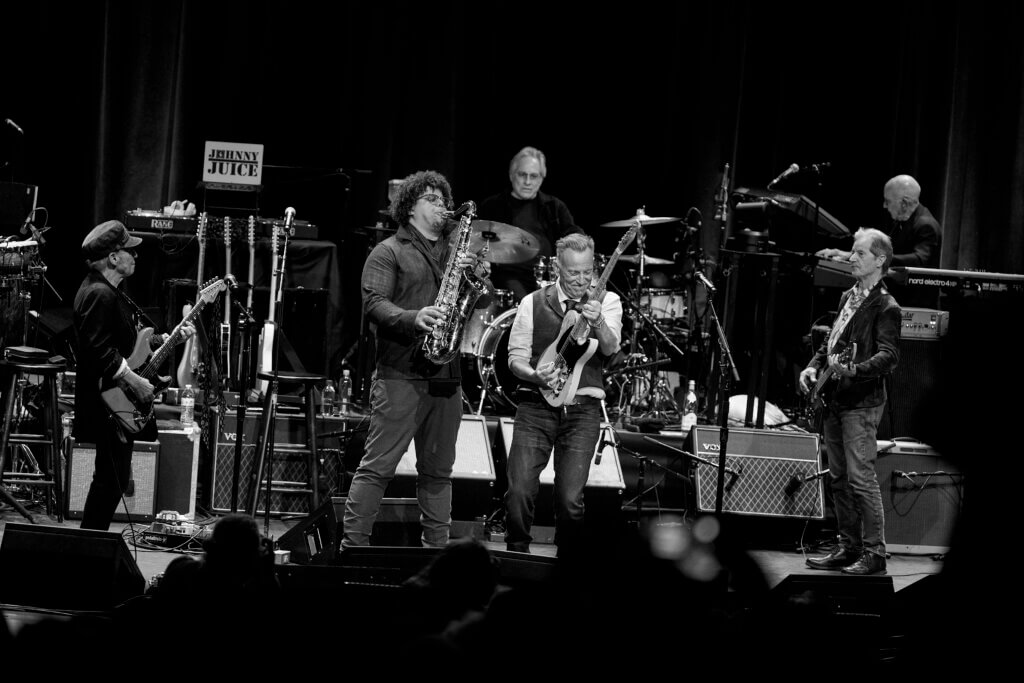 Black and white image of The E Street Band performing energetically on stage. Two musicians at center playing saxophones, flanked by guitarists, with a drummer and keyboardist in the background.