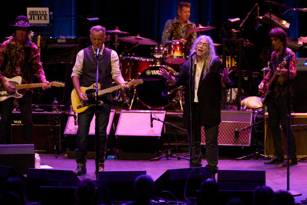 Bruce Springsteen and Patti Smith perform energetically on stage with guitars and drums, bathed in warm lighting. The scene captures a lively, vibrant concert atmosphere.