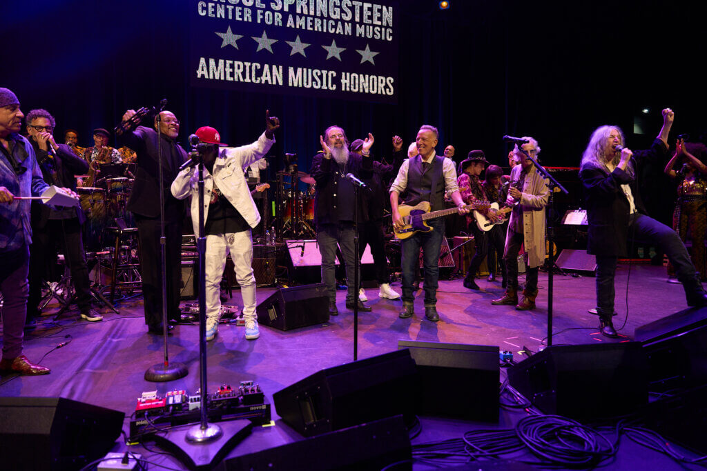 A lively stage performance with musicians and vocalists cheering and playing instruments. A banner reads "American Music Honors." The mood is celebratory.