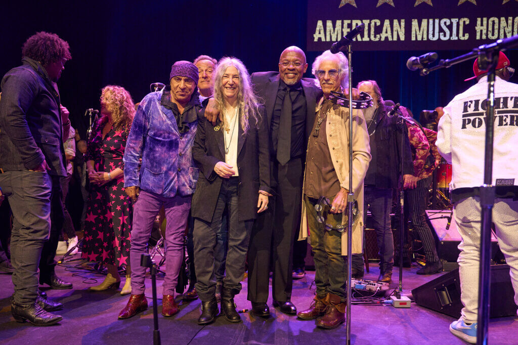 A diverse group of musicians and performers stand on stage at the "American Music Honors" event, exuding joy and celebration under bright stage lights. Stevie Van Zandt, Bruce Springsteen, Patti Smith, Dr. Dre and John Densmore
