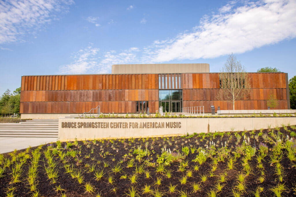 Exterior view of the Bruce Springsteen Center for American Music, featuring a modern building with a weathered steel facade, a glass entrance, and a concrete sign in the foreground surrounded by landscaped greenery under a blue sky.