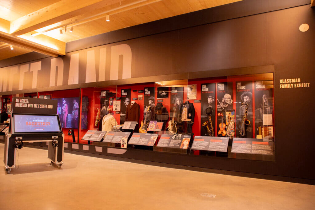 A museum exhibit titled "Glassman Family Exhibit" featuring a large, illuminated display case dedicated to the E Street Band, with individual sections for members including instruments like saxophones, violins, and guitars, alongside stage costumes and informational placards.