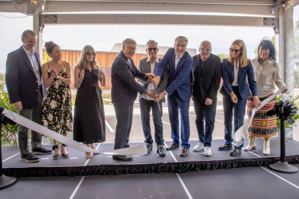 A diverse group of men and women smile while cutting a ribbon on a platform during an outdoor ceremony. The atmosphere is celebratory and formal. Including Melissa Kozlowski, Bob Santelli, Bruce Springsteen, President Pay Leahy, Jon Landau, Governor Sherrill and more.