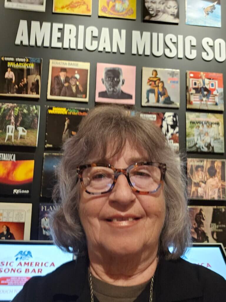 A woman with shoulder-length grey hair and tortoiseshell-framed glasses smiling at the camera. She is standing in front of a museum wall display titled "American Music," which features a grid of various vinyl record album covers.