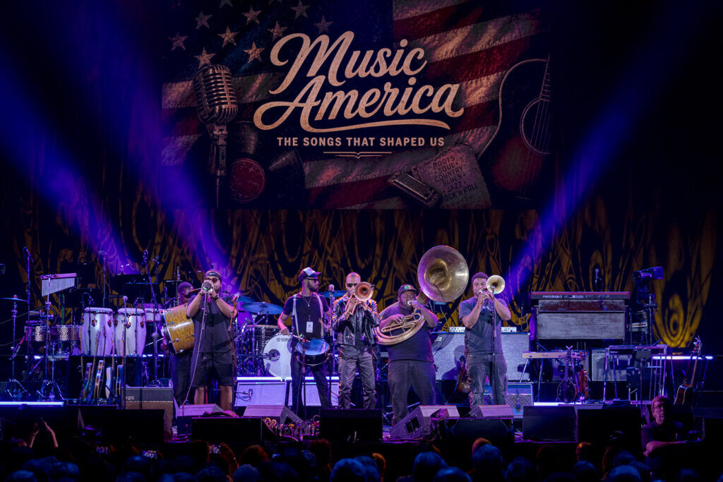 A lively brass band performs energetically on stage under purple spotlights. The backdrop reads "Music America: The Songs That Shaped Us."