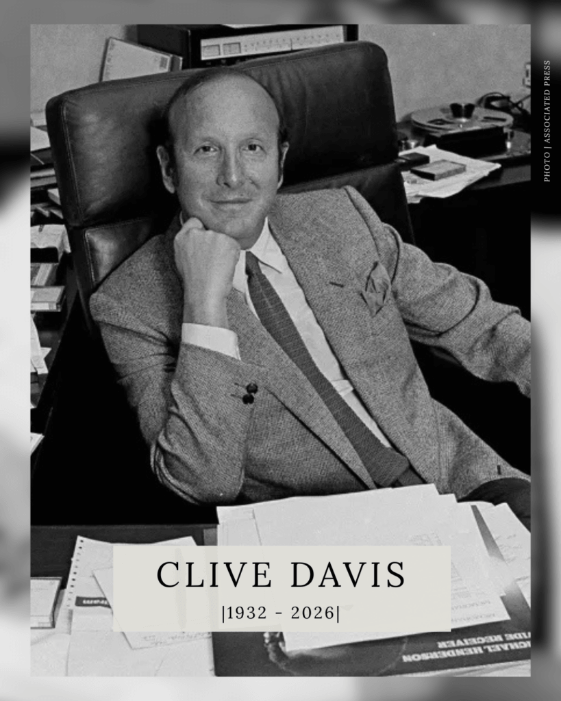 A black-and-white portrait of music executive Clive Davis sitting at a desk with papers, with a text overlay at the bottom that reads "CLIVE DAVIS |1932 - 2026|".