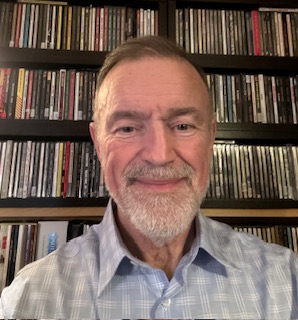 A portrait of a smiling, middle-aged man with short, thinning hair and a well-groomed grey beard, wearing a light blue and white plaid dress shirt. He is posed in front of several floor-to-ceiling shelves packed with a large collection of CD cases.