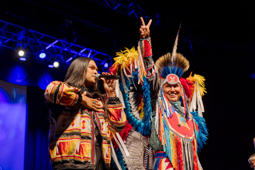 Two men are on stage, one singing into a microphone, both wearing vibrant, traditional Indigenous outfits. One raises a peace sign. They're under bright stage lights, exuding energy and pride.