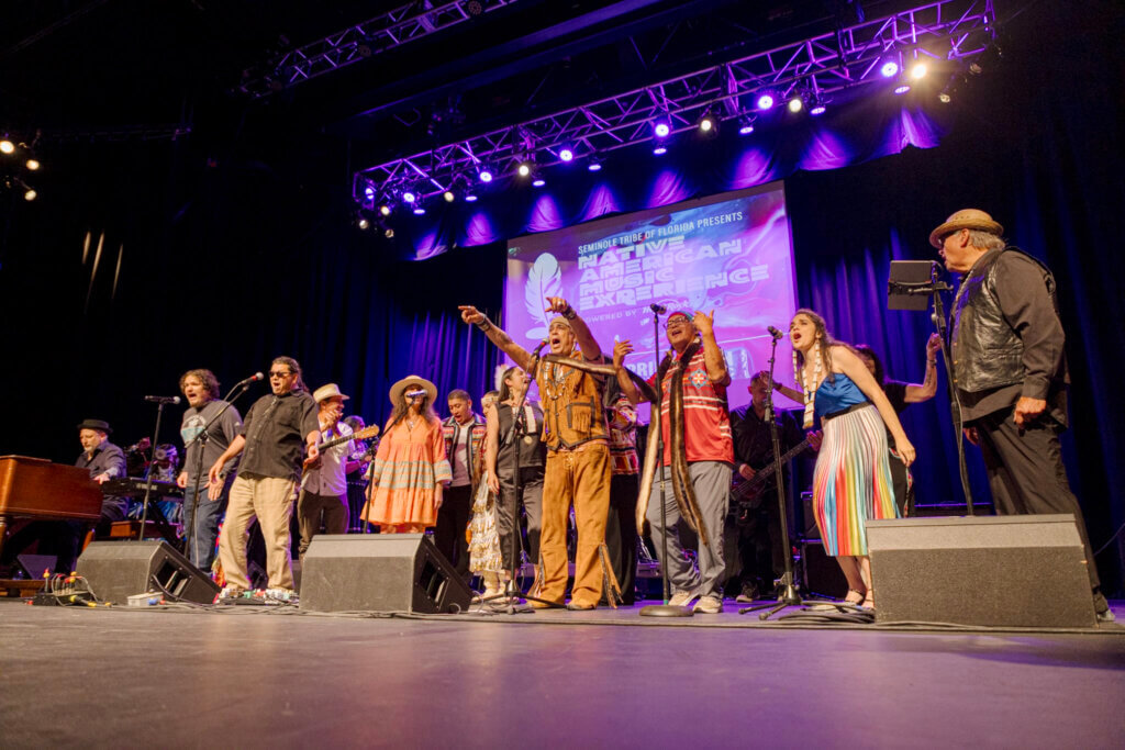 A diverse group of performers passionately sing on stage under vibrant purple lights. The backdrop displays a "Native American Experience" banner, creating an energetic and celebratory atmosphere.