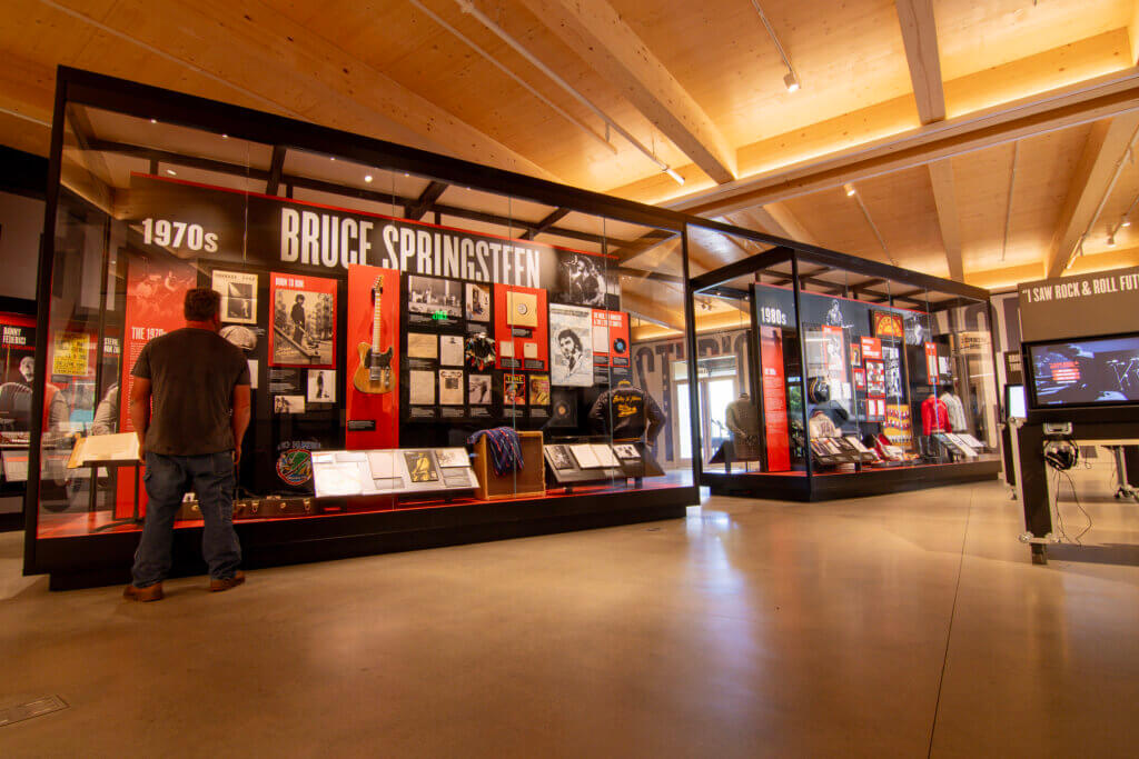 A museum visitor standing in front of a large, illuminated glass display case dedicated to Bruce Springsteen and the 1970s, filled with guitars, vinyl records, concert memorabilia, and vintage clothing.