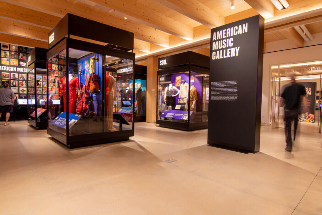Interior view of the American Music Gallery, featuring black display cases filled with musical artifacts and costumes, labeled with genre headings like "Blues" and "Soul," and a prominent sign reading "American Music Gallery."