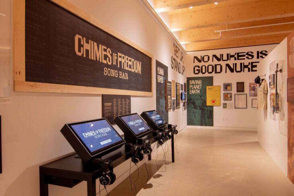 An exhibit titled "Chimes of Freedom Song Bar," featuring a wall with song titles, a row of interactive touch-screen kiosks with headphones, and surrounding wall displays with protest-themed posters and artifacts.