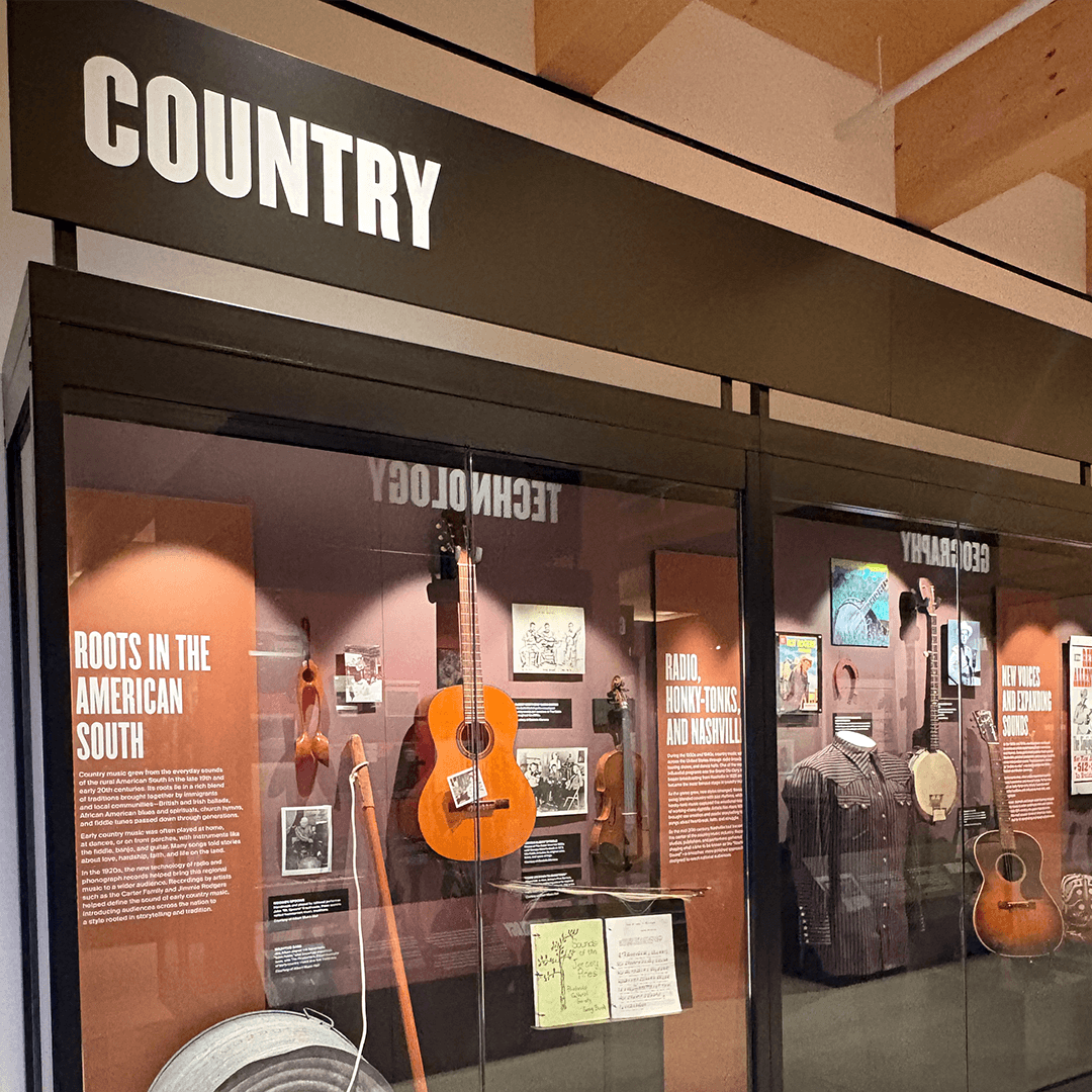 Museum exhibit themed "Country." Displays include a guitar, washboard, banjo, and various photos, highlighting the music's Southern roots. Warm lighting enhances a nostalgic tone.