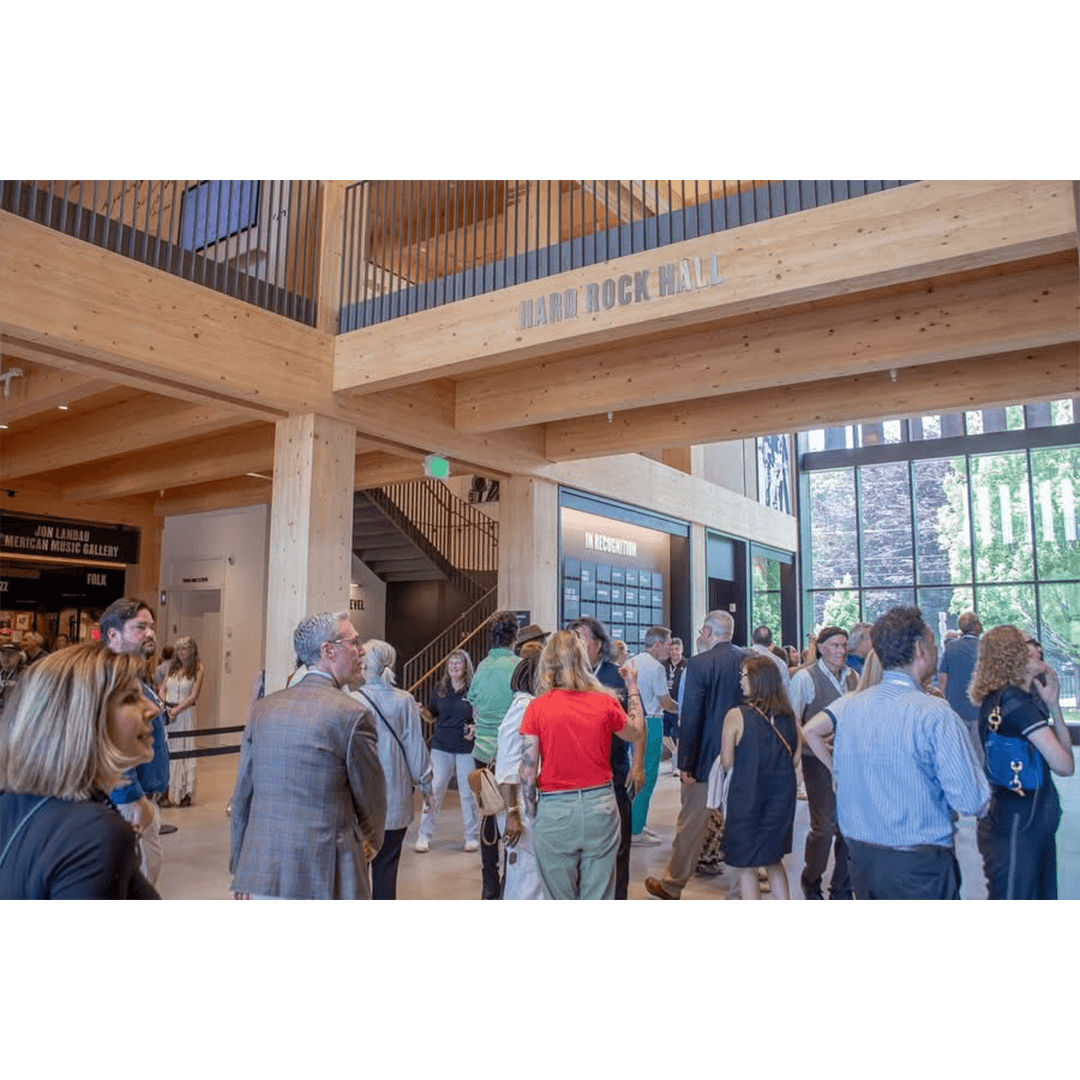 A photograph of a bustling event in the light-filled, wood-beamed interior of the Bruce Springsteen Center for American Music. A large group of people are socializing in the main hall, with a sign reading "Hard Rock Hall" visible on a prominent overhead beam, and signage for the "Jon Landau American Music Gallery" on the left.