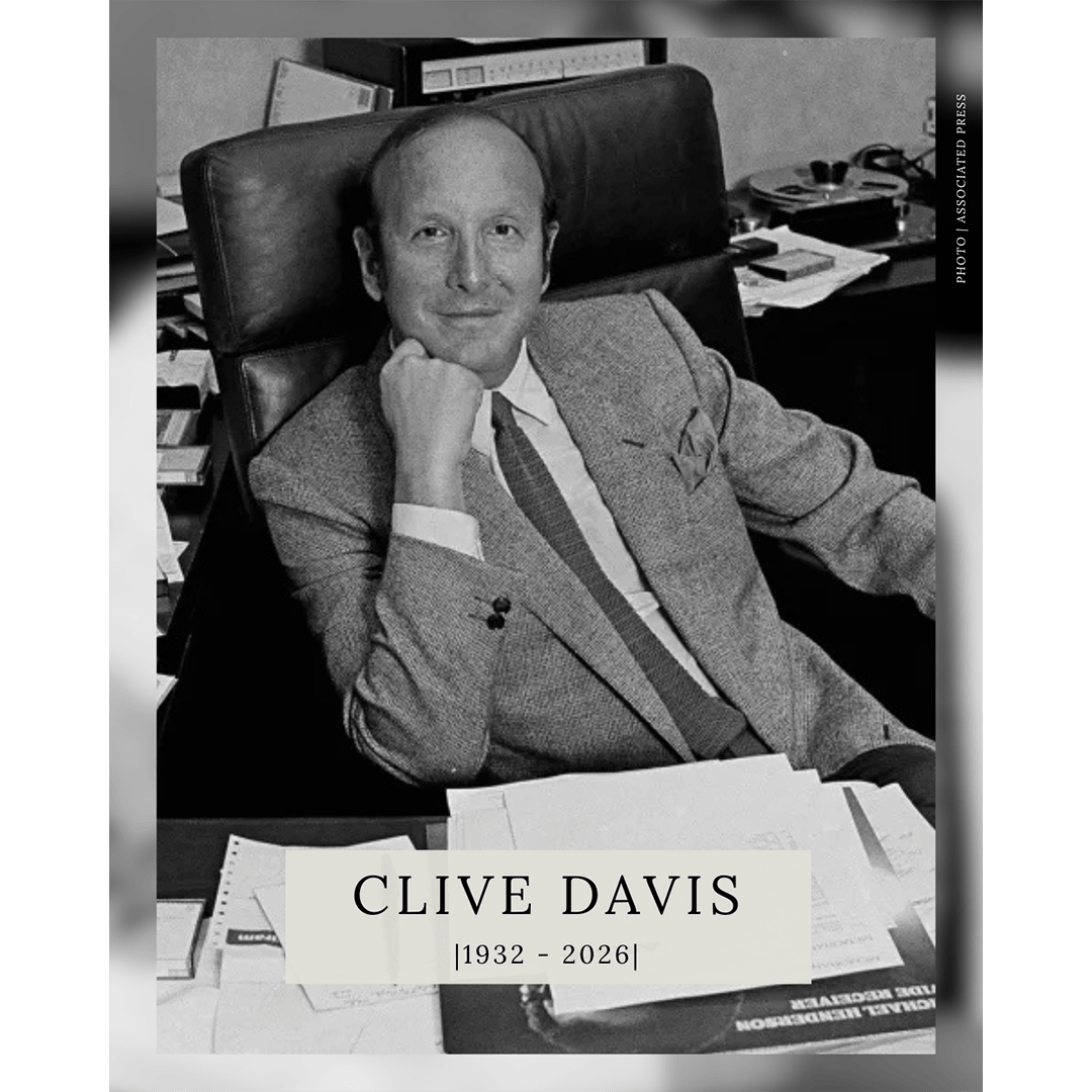 A black-and-white portrait of music executive Clive Davis sitting at a desk with papers, with a text overlay at the bottom that reads "CLIVE DAVIS |1932 - 2026|".